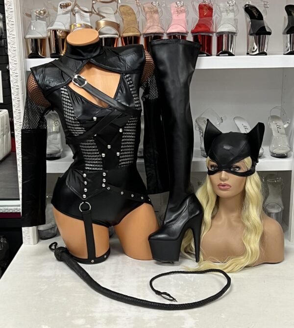 Front view of a glossy mannequin wearing a villainess bodysuit set with faux leather straps, mesh panels, O-ring garter, and zip back, posed beside black thigh-high platform boots and a cat ear mask wig display, with nude heels on shelves in the background.