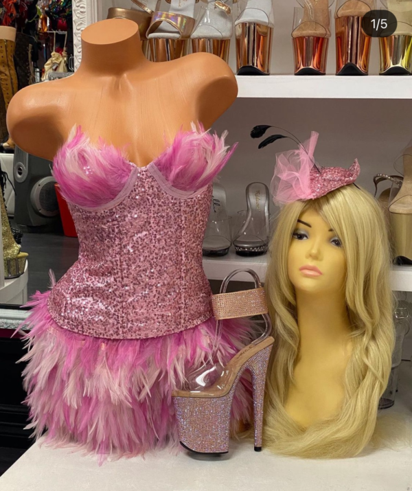 Front view of a glossy mannequin wearing a pink sequin corset with feather cups and a layered pink feather skirt, posed next to a glitter headband and pink rhinestone heels, displayed in a boutique with clear and rose gold platform heels in the background.
