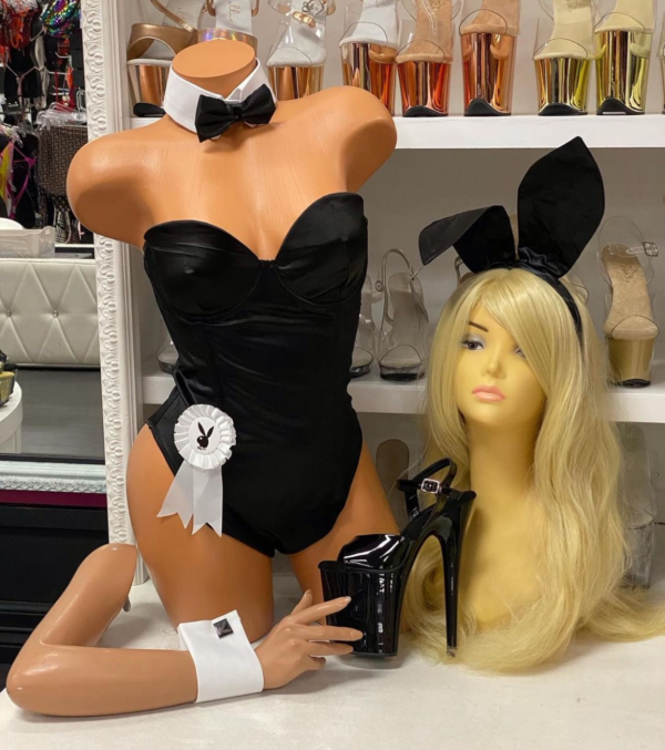 Front view of a glossy mannequin wearing a black satin corset bunny bodysuit with a white rosette and detachable collar and cuffs, posed next to a mannequin head in bunny ears and black patent heels, displayed in a boutique with platform shoes in nude and chrome finishes in the background.
