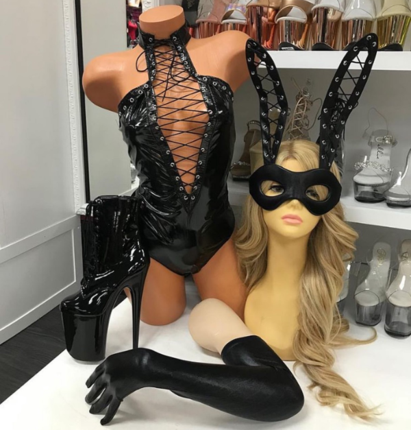 Front view of a glossy mannequin wearing a masked bunny costume with a black vinyl lace-up romper, long gloves, and lace-up bunny ears, posed next to patent black platform boots and a mannequin head with a mask, displayed in a boutique with chrome and rose-toned heels behind.
