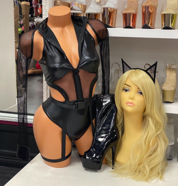 Front view of a glossy mannequin wearing a black cut-out kitty bodysuit with mesh sleeves, wet-look panels, belt, and leg garters, posed beside black patent platform booties and a blonde wig with cat ear headband, displayed in a boutique with shelves of nude and gold heels in the background.