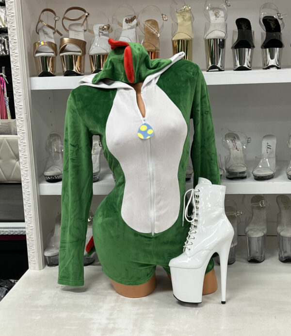Front view of a glossy mannequin wearing a green dino velvet romper with a white belly panel, red hood scales, plush stuffed tail, and novelty egg zipper pull, posed beside white lace-up platform heels, displayed in a boutique with shelves of clear and gold platform shoes in the background.