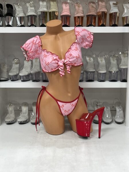 Front view of a glossy mannequin wearing a pink puff sleeve ruffle crop top and matching red-trimmed bow print thong bottoms, posed beside red patent platform heels, displayed in a boutique with clear and metallic heels in the background.