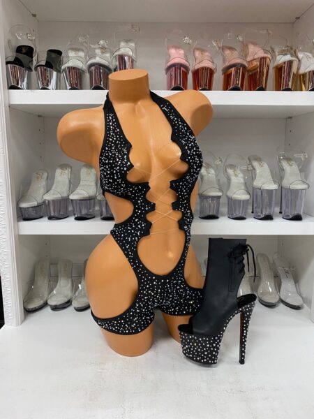 Front view of a glossy mannequin wearing a black rhinestone romper with sculpted cutouts and center lace-up front, posed next to a black peep-toe platform boot with rhinestones, displayed in a boutique with shelves of high heels in the background.