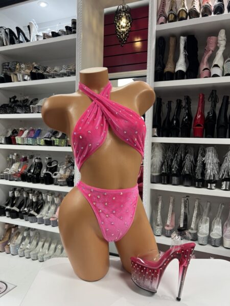 Front view of a glossy mannequin wearing a hot pink crisscross velvet romper with pearl rhinestones, posed next to ombré clear and pink platform heels, displayed in a boutique with shelves of sparkling heels in the background.