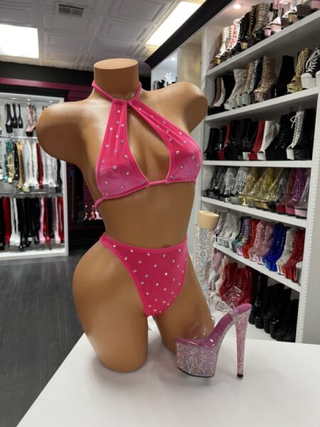 Front view of a glossy mannequin wearing a hot pink rhinestone bikini set with a keyhole halter top, posed next to clear rhinestone platform heels, displayed in a boutique with shelves of glittery high heels in the background.