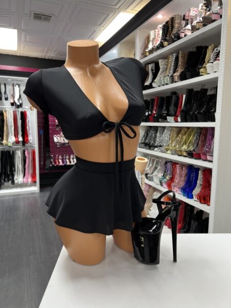 Front view of a glossy mannequin wearing a black satin-look tie-front cap-sleeve crop top with a matching high-waisted flared mini skirt, posed next to black patent platform heels with ankle straps, displayed in a boutique with shelves of rhinestone and patent platform boots in the background.