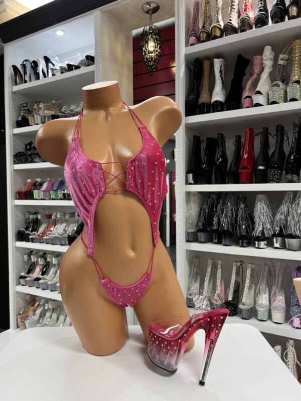 Front view of a glossy mannequin wearing a hot pink velvet cut-out romper with rhinestones and lace-up detail, posed next to ombré pink rhinestone high heels, displayed in a boutique with black, red, white, and glitter platform boots in the background.