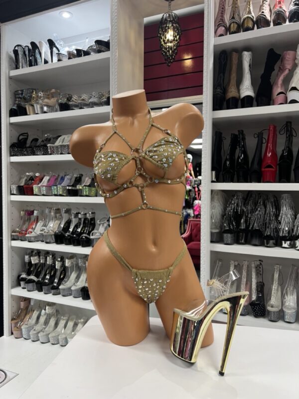 Front view of a glossy mannequin wearing a gold rhinestone bikini with triangle crystal top, halter straps, and matching cutout velvet bottom, posed next to gold chrome platform heels, displayed in a boutique with heels and boots in the background.