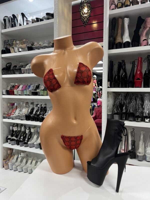 Front view of a glossy mannequin wearing a red micro bikini with black logo print, posed next to matte black lace-up platform stiletto boots, displayed in a boutique with shelves of rhinestone heels and patent boots in the background.