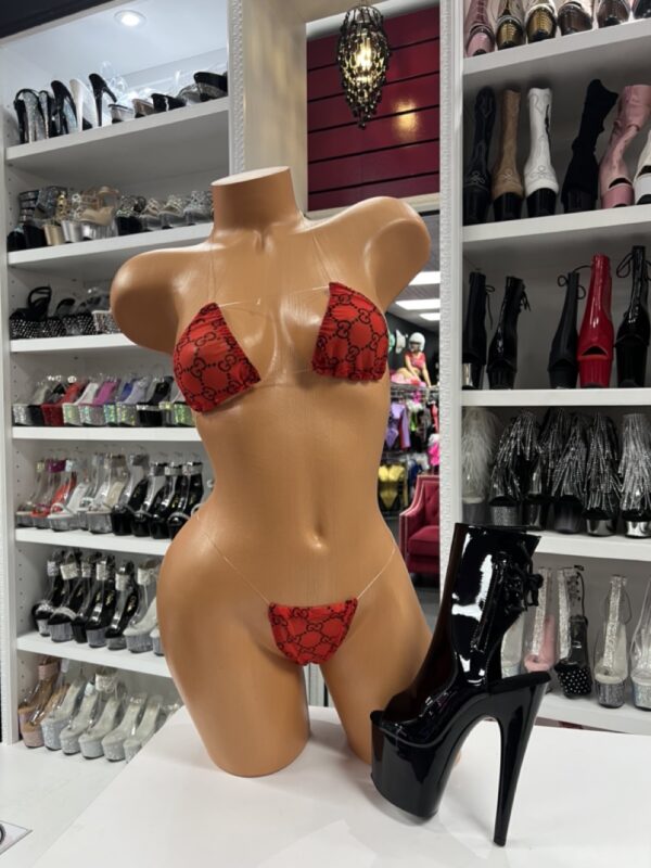 Front view of a glossy mannequin wearing a red micro bikini with black logo print, posed next to black patent platform lace-up boots, displayed in a boutique with shelves of high heels in the background.