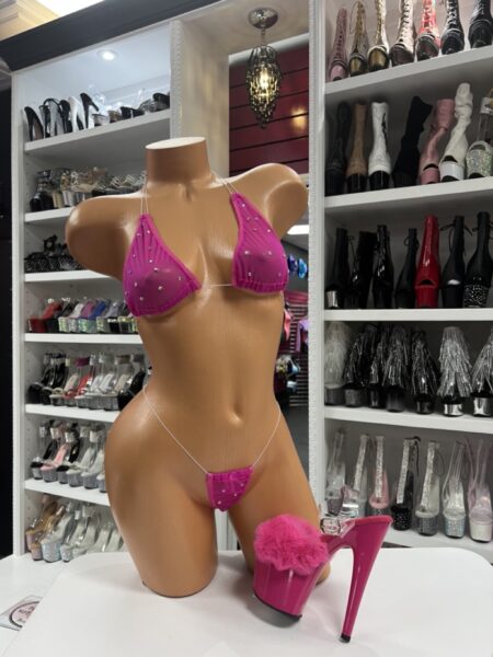 Front view of a glossy mannequin wearing a hot pink micro bikini with rhinestone detailing and clear straps, styled next to a hot pink marabou heel, displayed in a boutique with shelves of clear, silver, and red platform boots in the background.