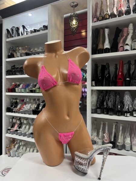 Front view of a glossy mannequin wearing a neon pink micro bikini with rhinestones and ruffle trim, featuring clear halter and hip straps, posed next to clear platform heels with silver studs, displayed in a boutique with shelves of black and silver boots in the background.