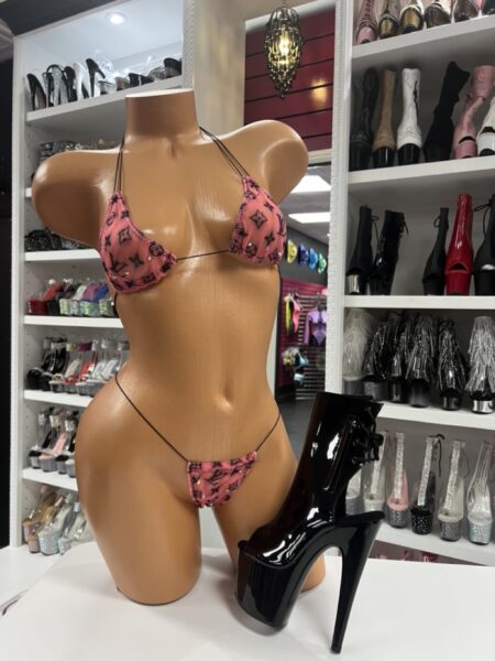 Front view of a glossy mannequin wearing a coral mesh designer-inspired micro bikini with black straps, posed next to black patent lace-up platform boots, displayed in a boutique with shelves of rhinestone and clear heels in the background.