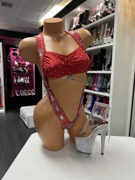 Front view of a glossy mannequin wearing a red rhinestone-embellished bandeau bodysuit with twisted top and crystal straps, posed next to a silver glitter platform heel, displayed in a boutique with shelves of colorful performance heels in the background.